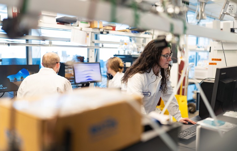 A woman operates assay equipment in a lab, as two other people work behind her.