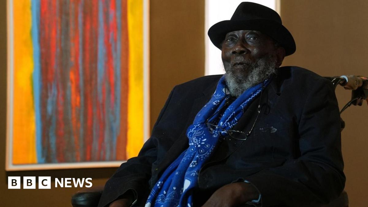 Sir Frank Bowling looks at the camera unsmiling. He is in a wheelchair and behind him is a brightly coloured orange and red abstract painting. Sir Frank is wearing a black hat and jacket and blue scarf.