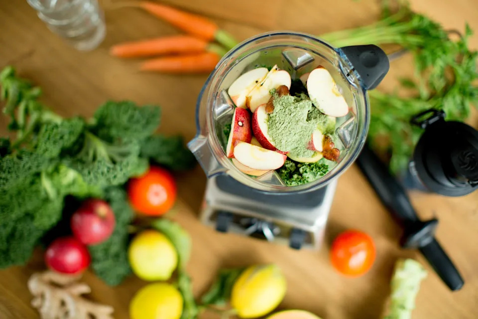 Blender filled with apple slices, leafy greens, and powder sits on a counter with fresh vegetables and fruits nearby