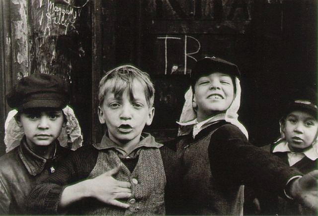 Four young boys stand outside a worn building, playfully posing with confident expressions. Graffiti is visible on the wall behind them