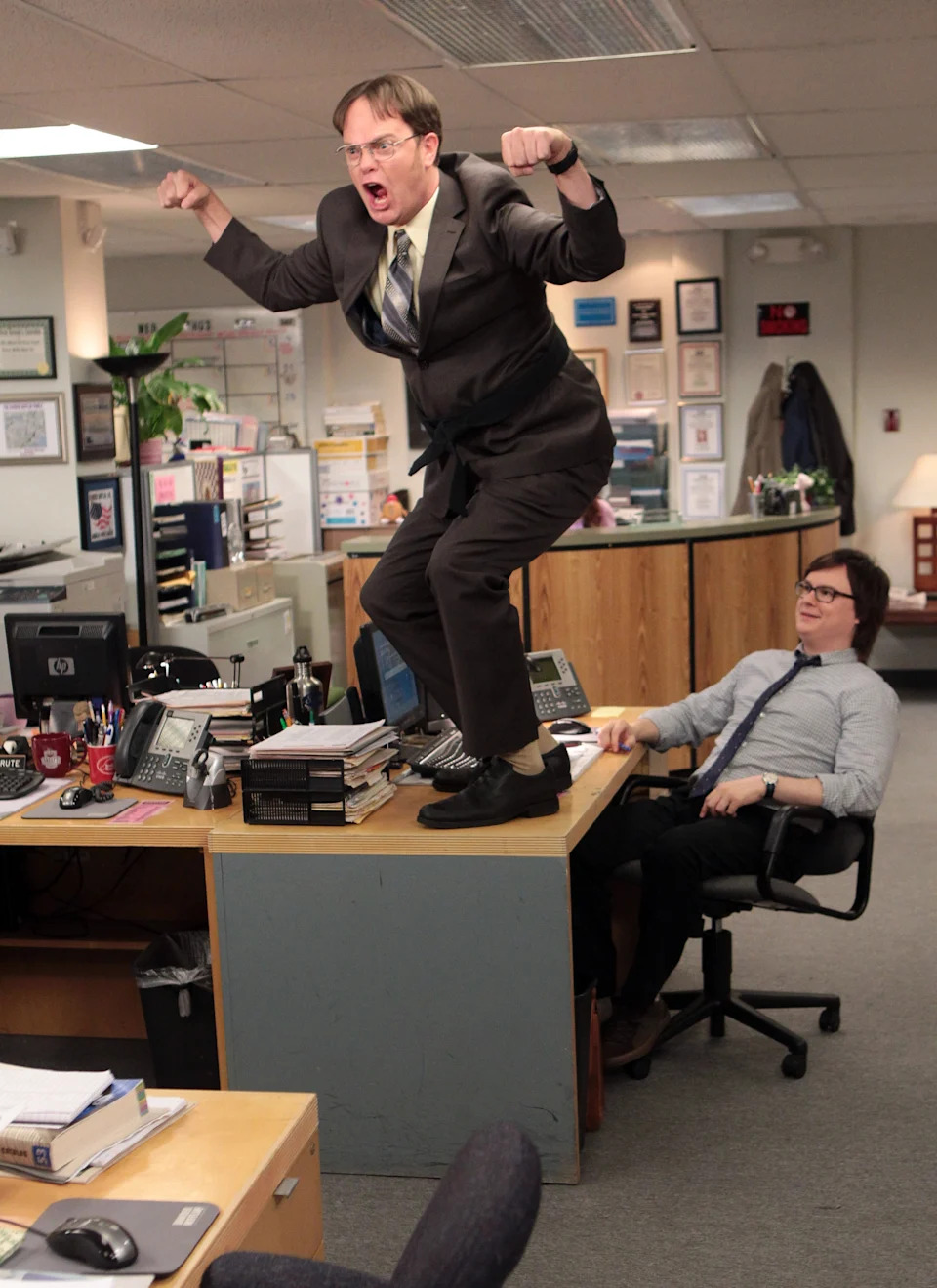 A man in a suit stands and poses energetically on a desk in an office, while a seated colleague watches with mild surprise