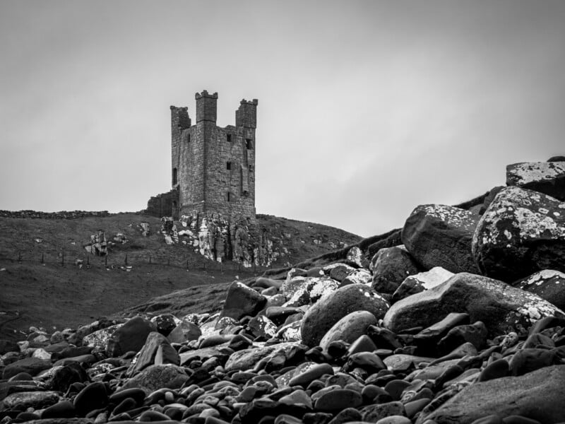 A black and white photo of a ruined stone castle tower on a grassy hill, with large rocky boulders in the foreground under a cloudy sky.