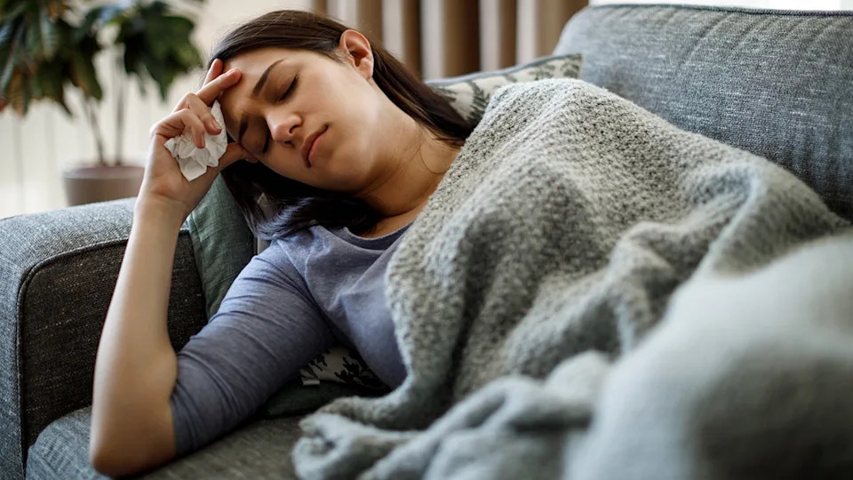 Woman resting on a couch with a blanket, holding a tissue to her head and appearing fatigued at home.