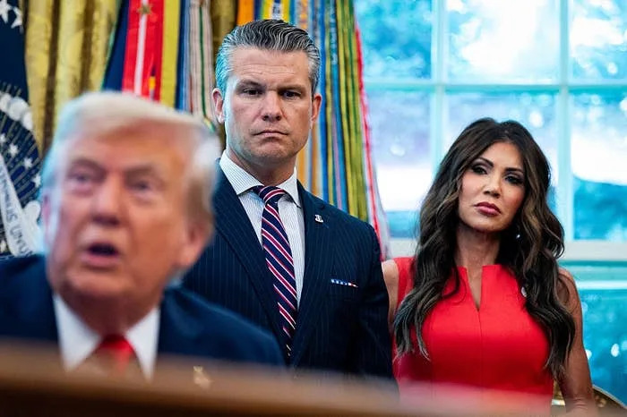 Three people are in a room, one speaking in the foreground, with two listening intently in the background