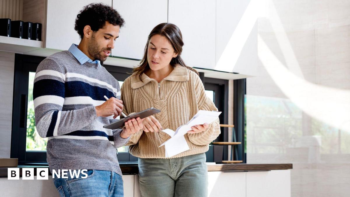 Couple in a kitchen look at a tablet and some paperwork.