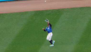 Byron Buxton, wearing a light blue uniform, white pants, and black socks, reaches up with his glove to make a catch in left center field.
