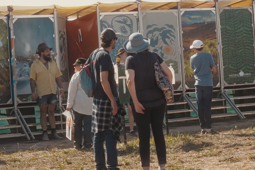 A row of composting toilets with colourful external doors at a festival, with people using them.