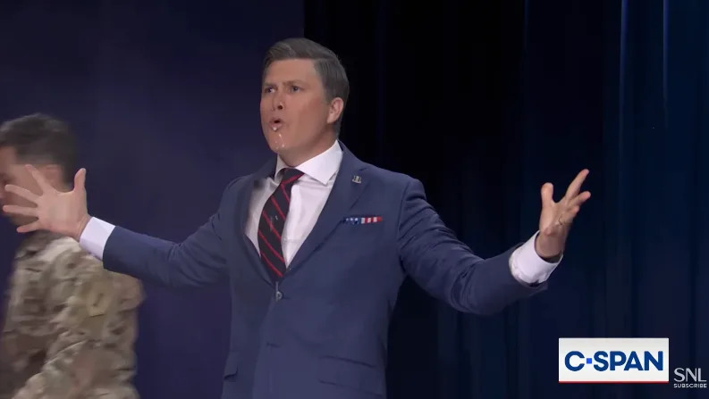 Person in a suit on stage, gesturing with arms wide. C-SPAN and SNL logos in corner
