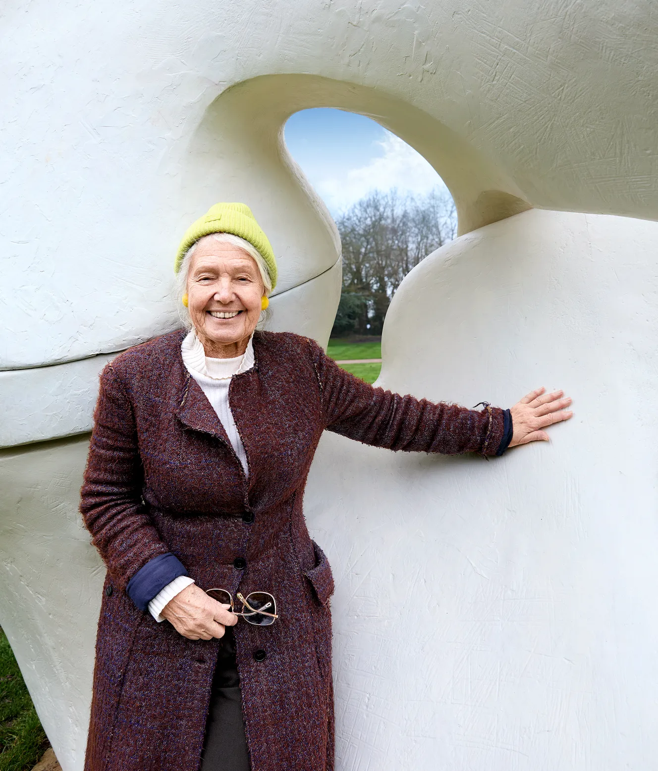 Mary Moore, daughter of sculptor Henry Moore, stands next to a large abstract white sculpture with a hole, smiling at the camera.