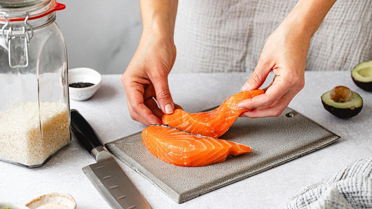 Woman's hands seen prepping raw salmon on cutting board with rice, knife and avocado around her.