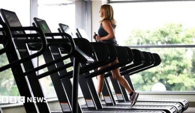 A woman wearing gym gear runs of a treadmill near large glass windows with trees outside. There are several empty treadmills next to her.