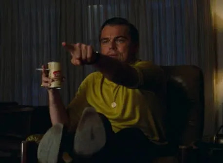 Person sitting in a chair, wearing a casual outfit, holding a drink and cigarette, gesturing forward with focus