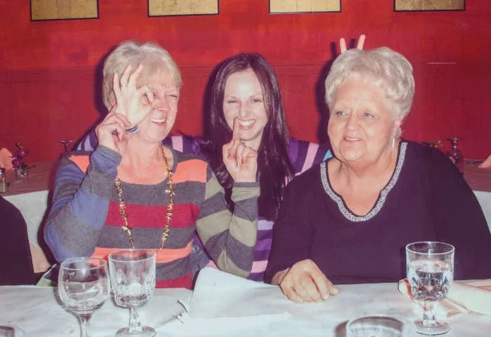 Three women smiling at a dining table, two making playful gestures with their hands. Glasses and plates are set on the table in front of them