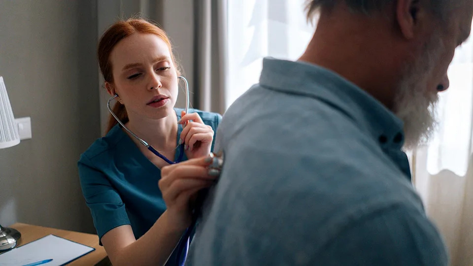 Nurse listening with stethoscope to back of mature adult man in hospital room.