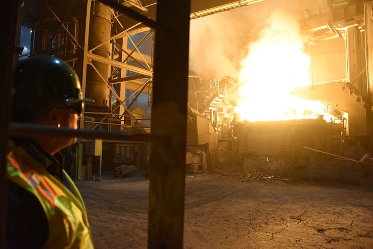 A man in a hard hat and reflective vest looks at an industrial fire.