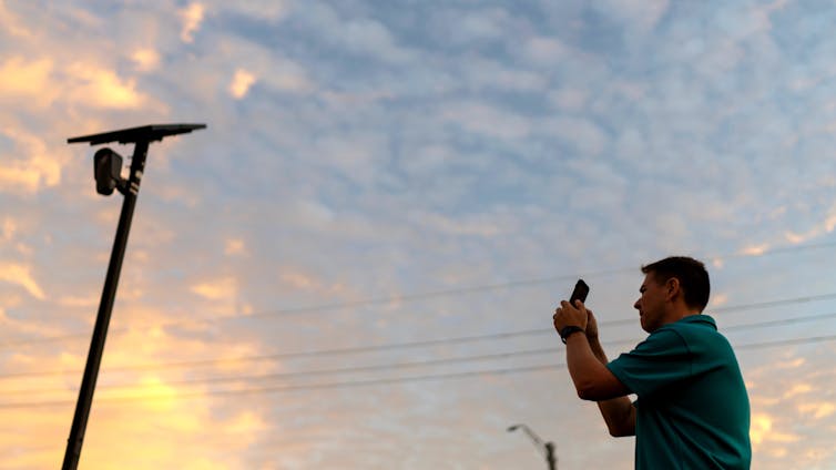 Young man wearing a polo shirt uses his smartphone to take a photograph of a camera affixed to a poll at dusk.