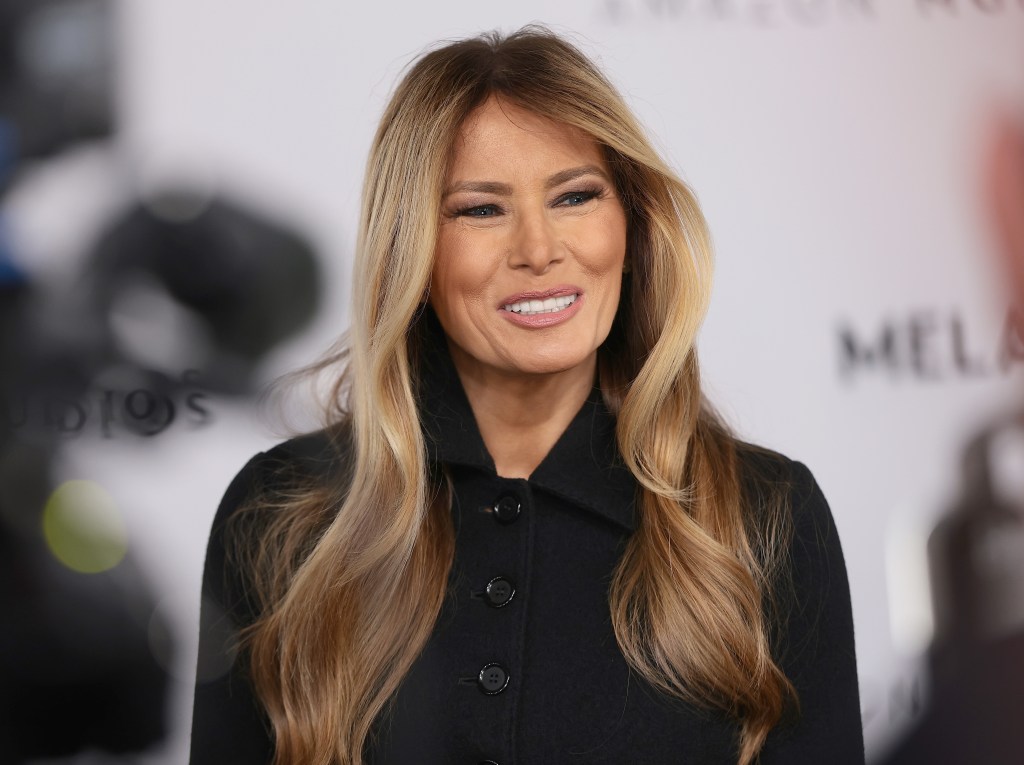 Melania Trump, wearing a black coat, smiles slightly.