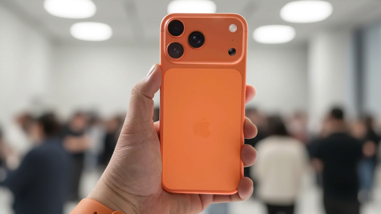 A Cosmic Orange iPhone 17 Pro Max held up infront of a blurry crowd of people in an individual's left hand
