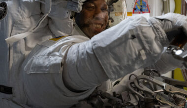 Chris Williams, an astronaut, is wearing a white spacesuit and helmet. He is facing the camera, but his body is turned to the right of the image. His arms are extended in front of him.