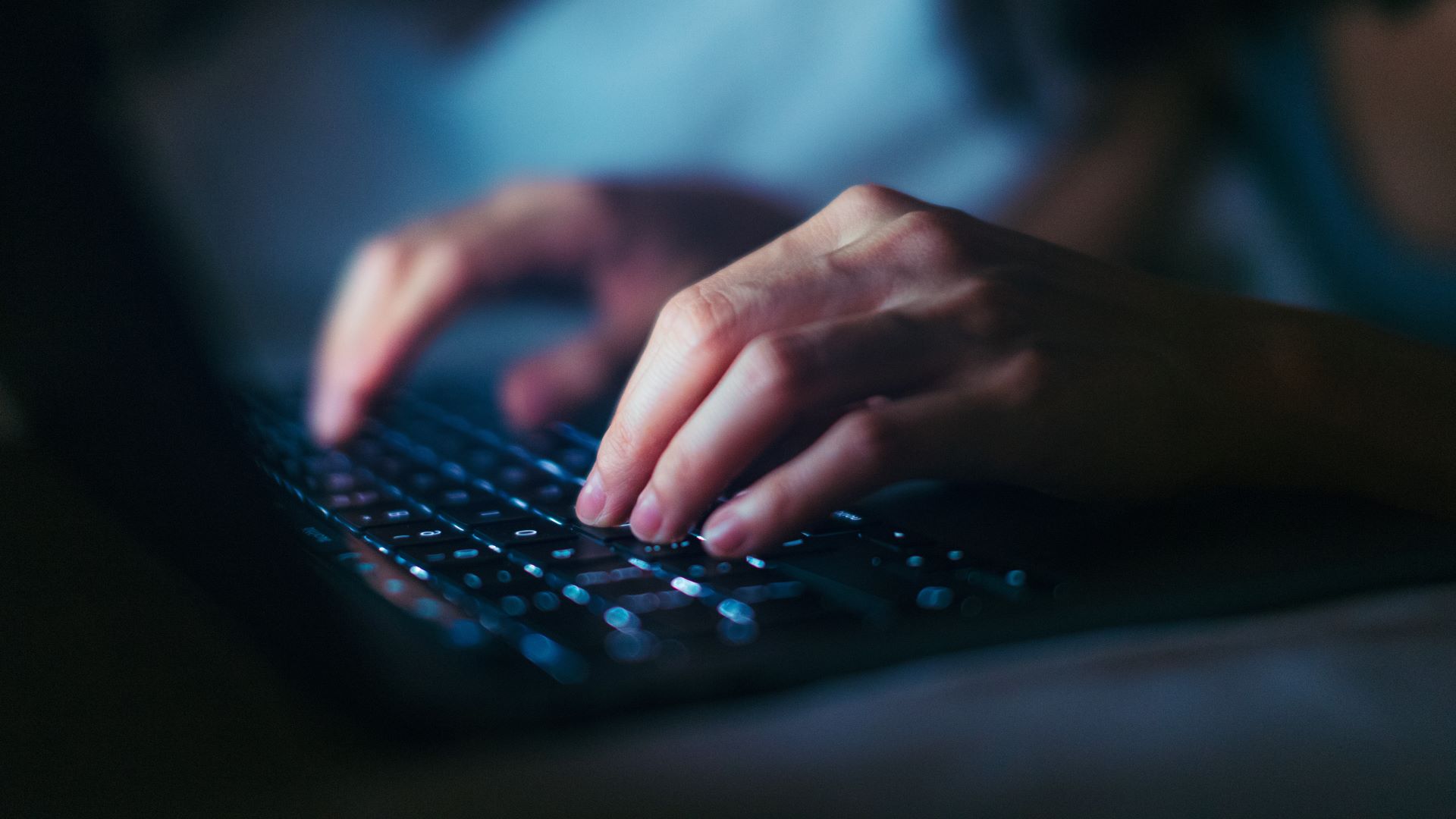 A close-up photograph of a person's hands typing on a backlit laptop keyboard