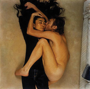 A nude person curls up and kisses a clothed person on a beige surface