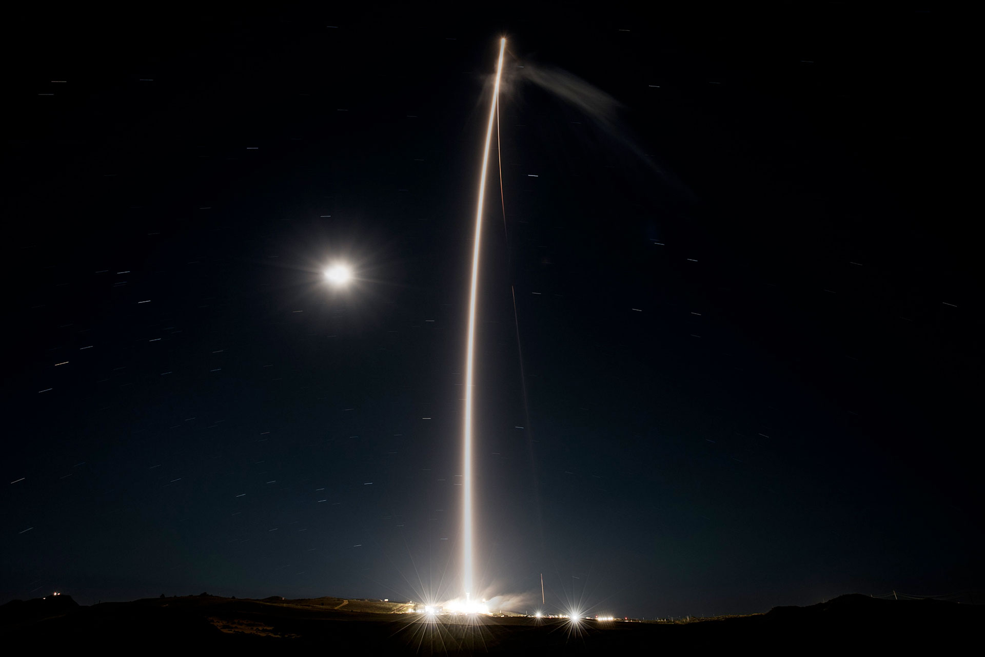 a time lapse photo captures the glowing streak of a rocket's plume as it climbs spaceward adding to the light from the moon