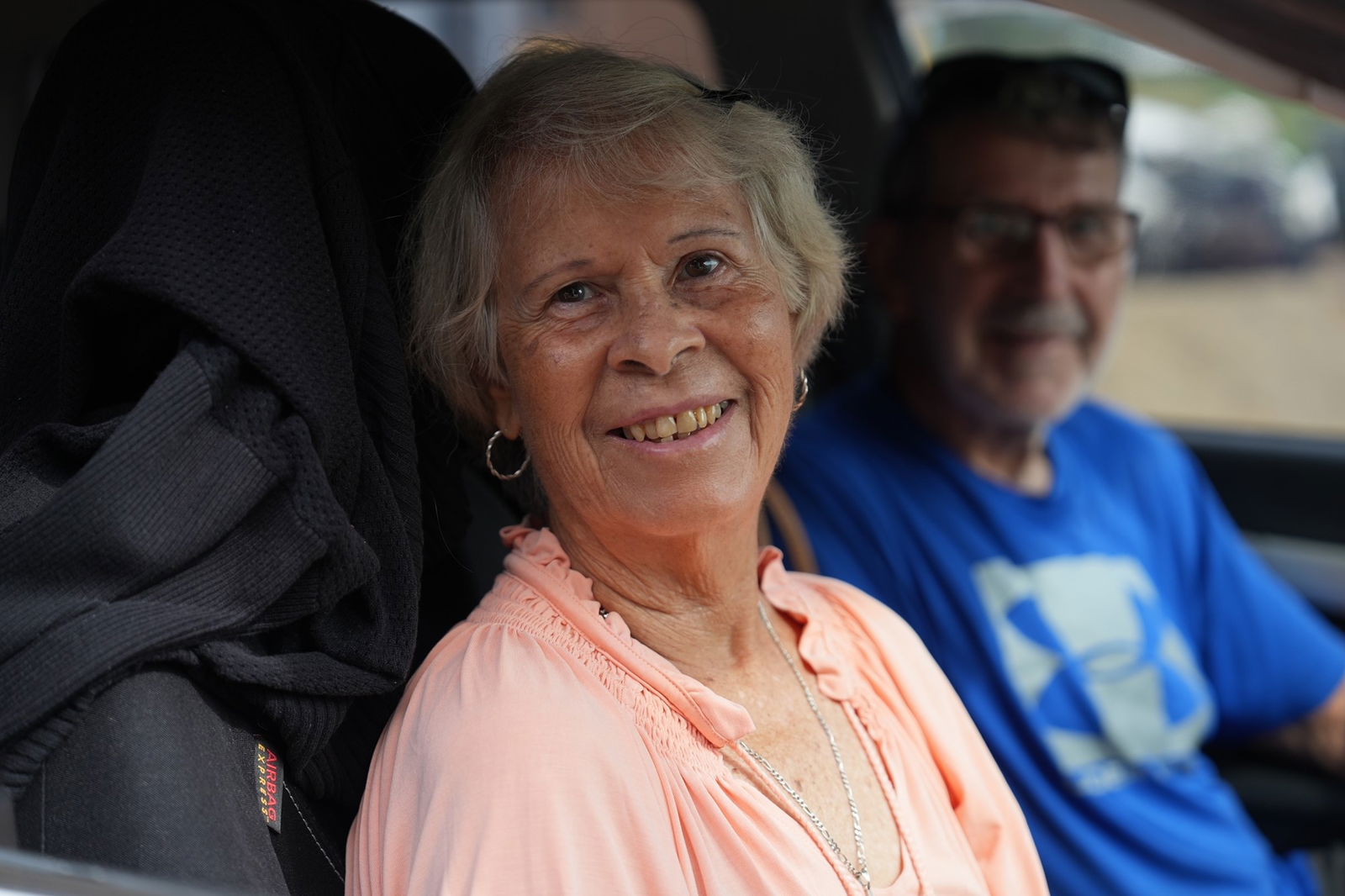 A woma with short gray hair, tanned complexion, wearing a peach blouse, sitting in a car.