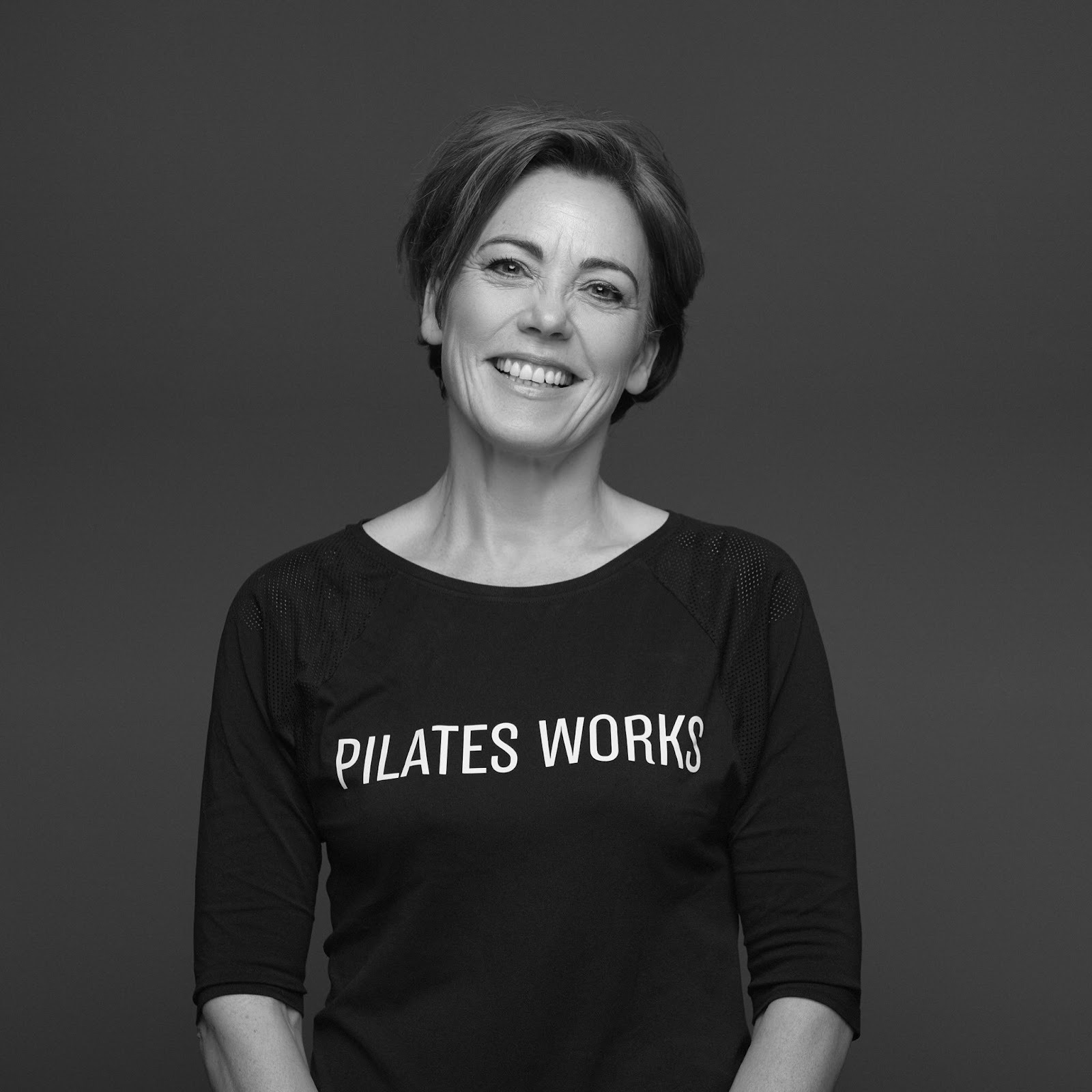 Woman, wearing black T-shirt with “Pilates Works” written on it in white text, smiles at camera