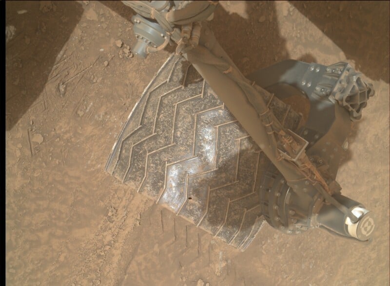 Close-up view of a Mars rover wheel with a chevron tread pattern on the dusty, rocky Martian surface. Mechanical parts of the rover’s suspension and arm are visible above the wheel.