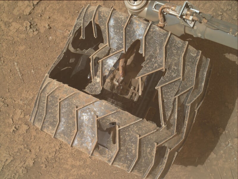 A close-up view of a damaged rover wheel on a dusty, rocky surface. The metallic wheel shows significant cracks, holes, and deformation along its treads, revealing internal mechanical components.