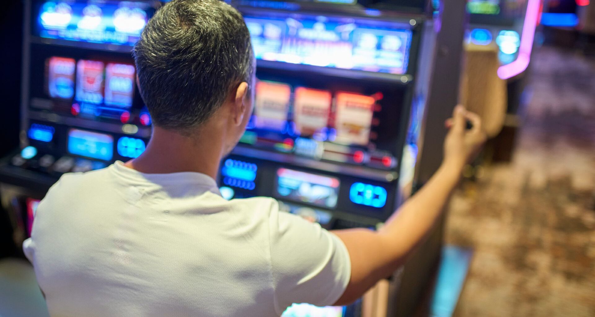A view over the shoulder of a man wearing a white t-shirt with gray hair sitting at a slot machine