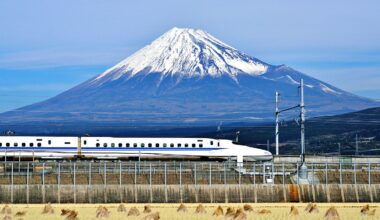 A white bullet train speeds along tracks in the foreground, with Japan’s snow-capped Mount Fuji towering in the background under a clear blue sky.