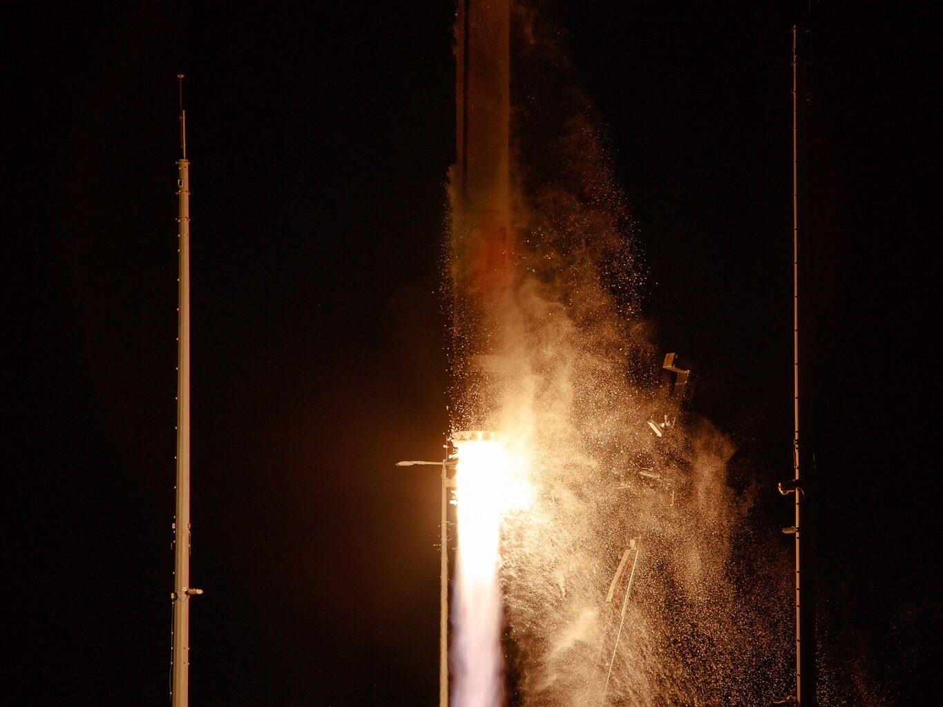 A Rocket Lab HASTE rocket lifts off at night from NASA's Wallops Island, with bright orange and white flames illuminating billowing clouds of smoke against a black sky. Launch structure towers are visible on either side of the rocket.