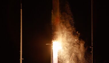 A Rocket Lab HASTE rocket lifts off at night from NASA's Wallops Island, with bright orange and white flames illuminating billowing clouds of smoke against a black sky. Launch structure towers are visible on either side of the rocket.