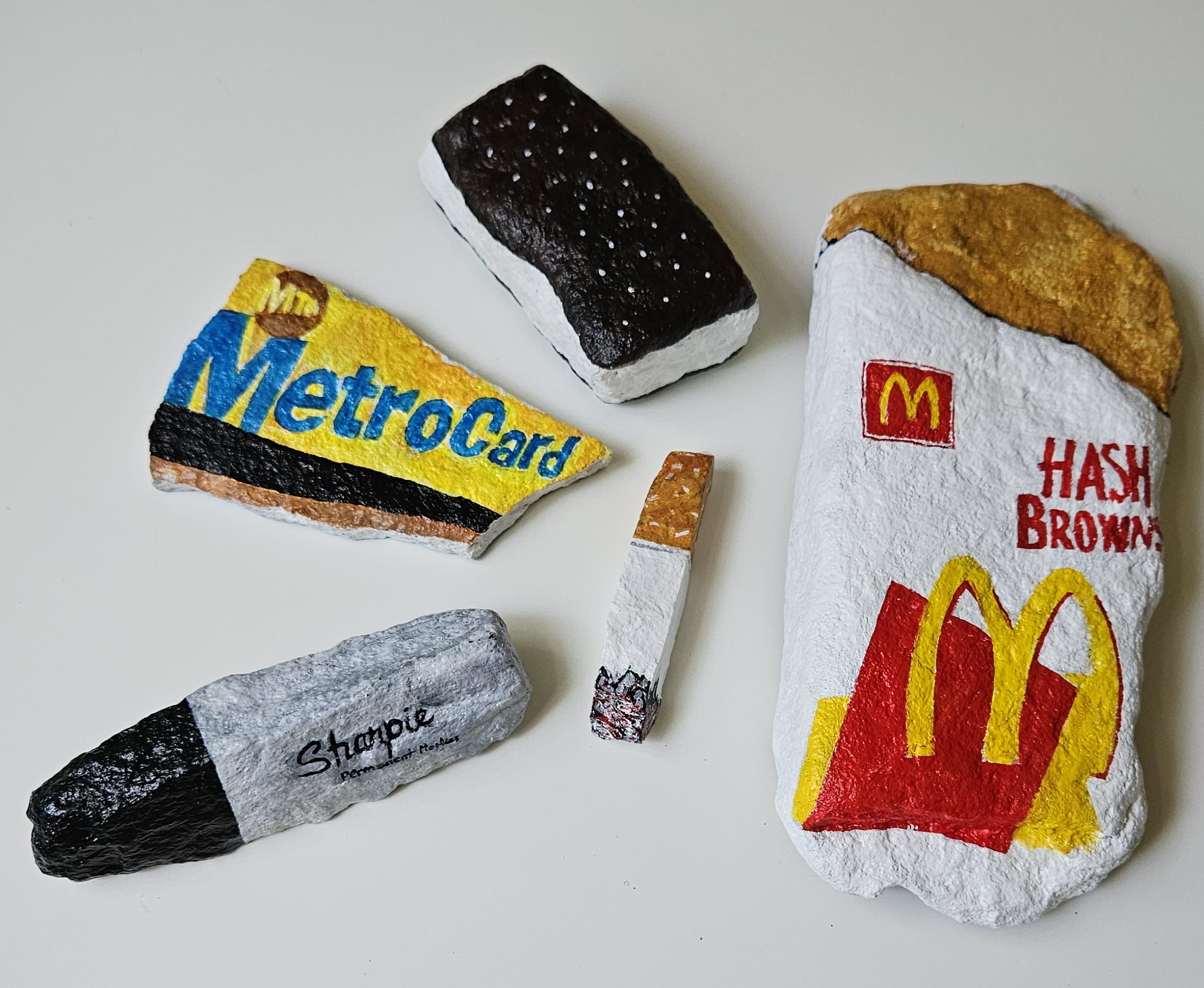 Painted rocks by Elizabeth Saloka that resemble a Metro card, Sharpie marker, McDonald's hashbrown, an ice cream sandwich, and a cigarette