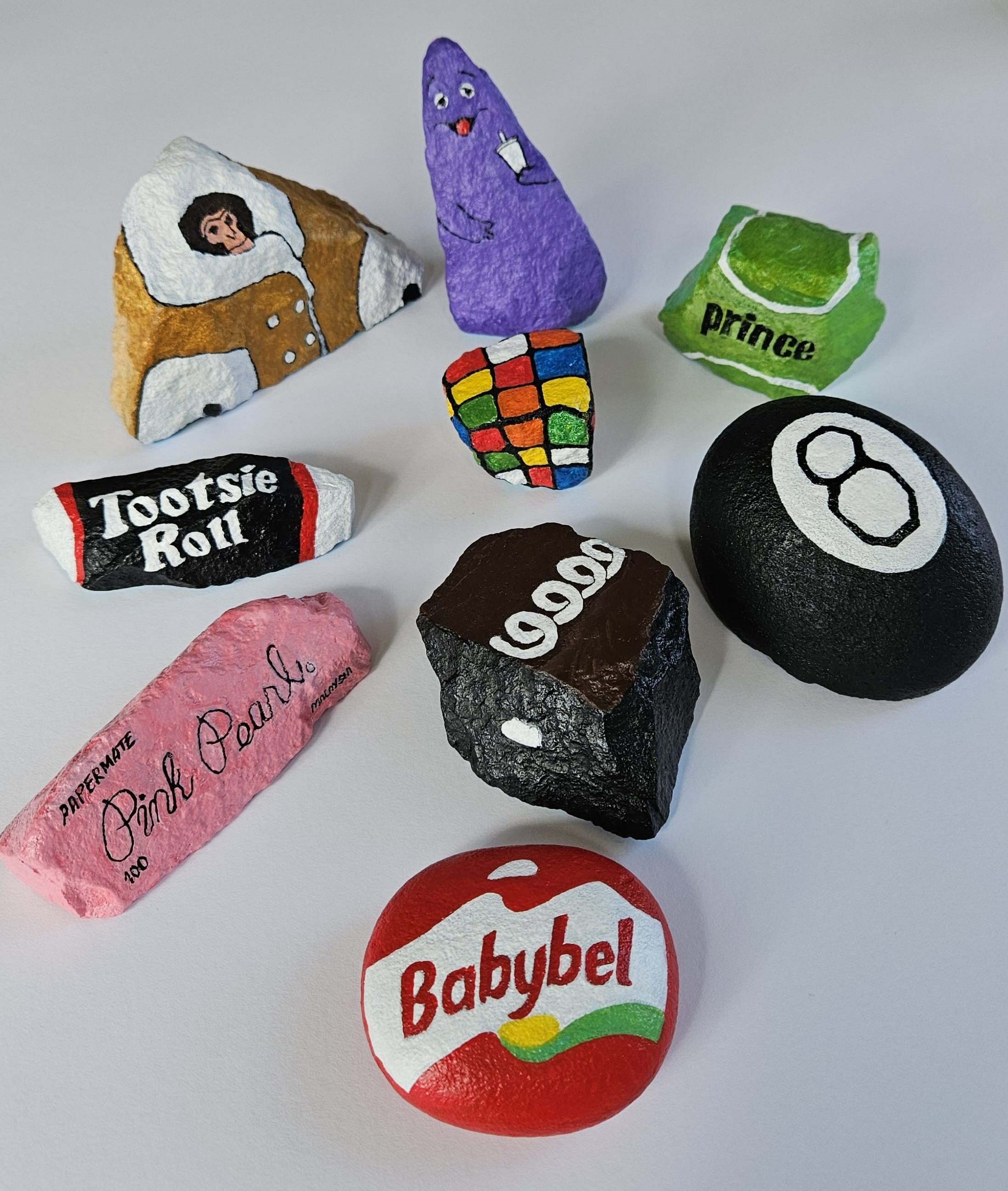 Painted rocks by Elizabeth Saloka that resemble objects like a Babybel cheese round, a Tootsie Roll, a Hostess cake, an 8 ball, a Rubik's cube, and other objects