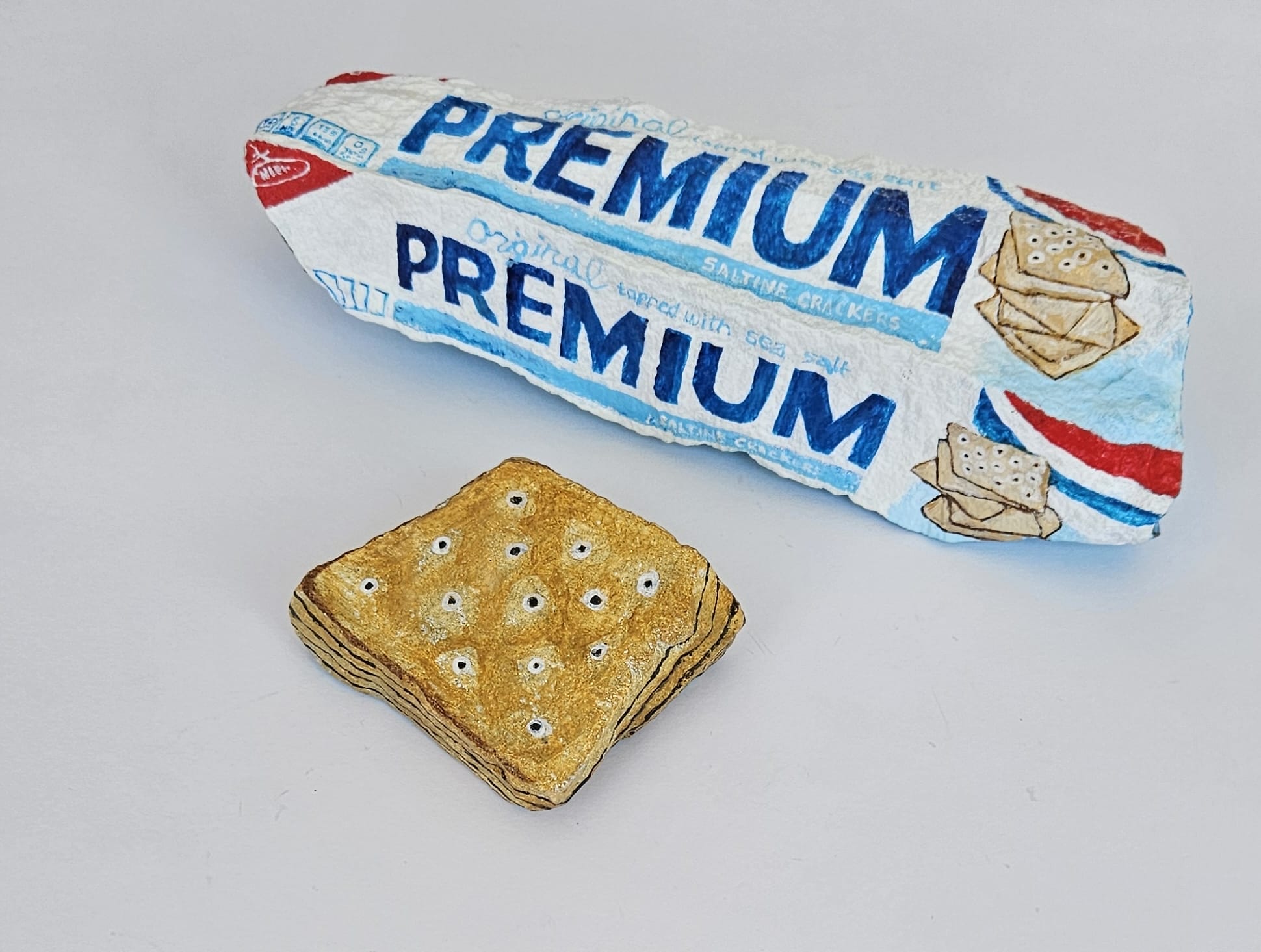 Painted rocks by Elizabeth Saloka that resemble a box of Premium saltine crackers and a single saltine cracker on its own