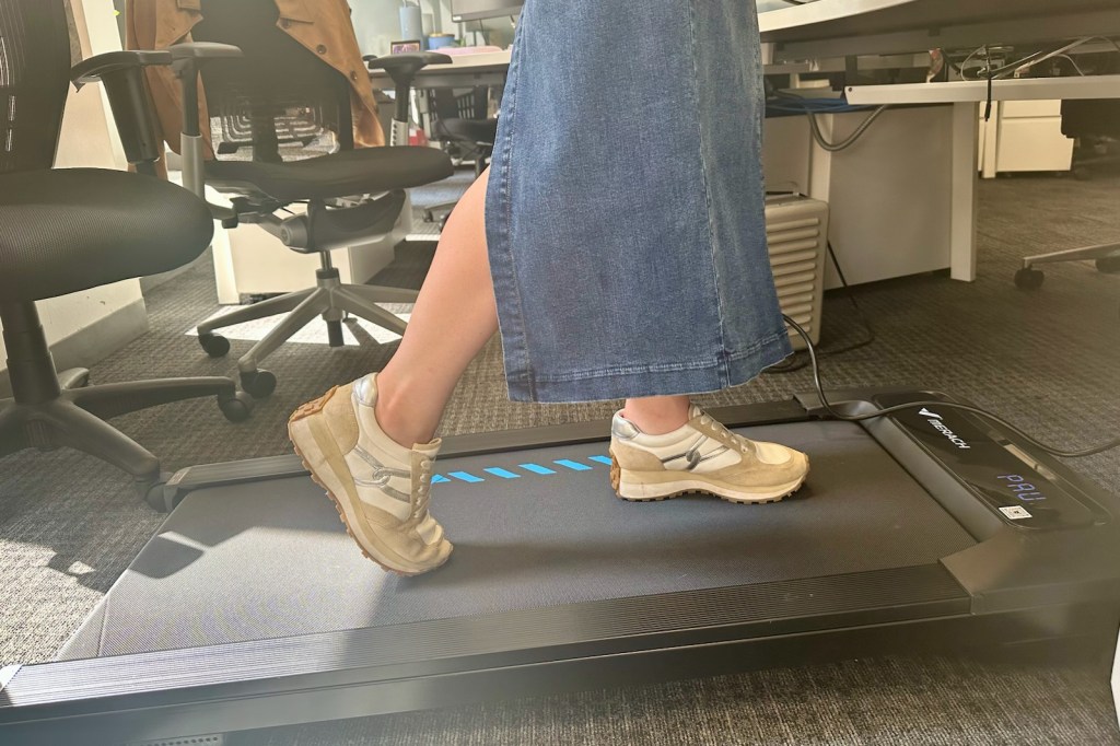 A person in a denim skirt and sneakers walks on a compact treadmill in an office setting.