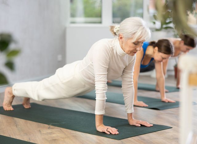 Concentrated elderly woman holding plank pose to strengthen body muscles during group yoga training in studio. Core exercises for older adults. Active lifestyle concept..
