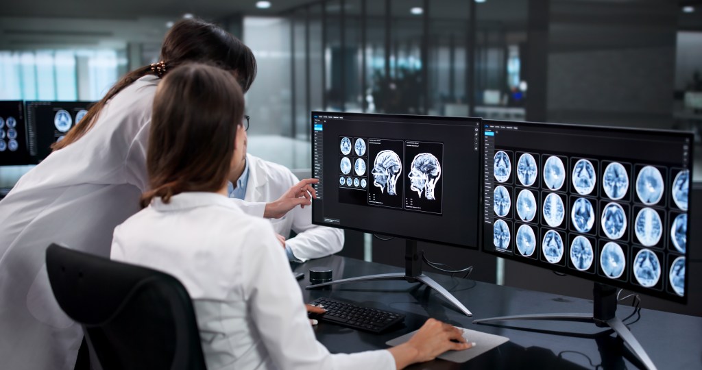 Three neurologists analyze brain scans on multiple computer monitors.