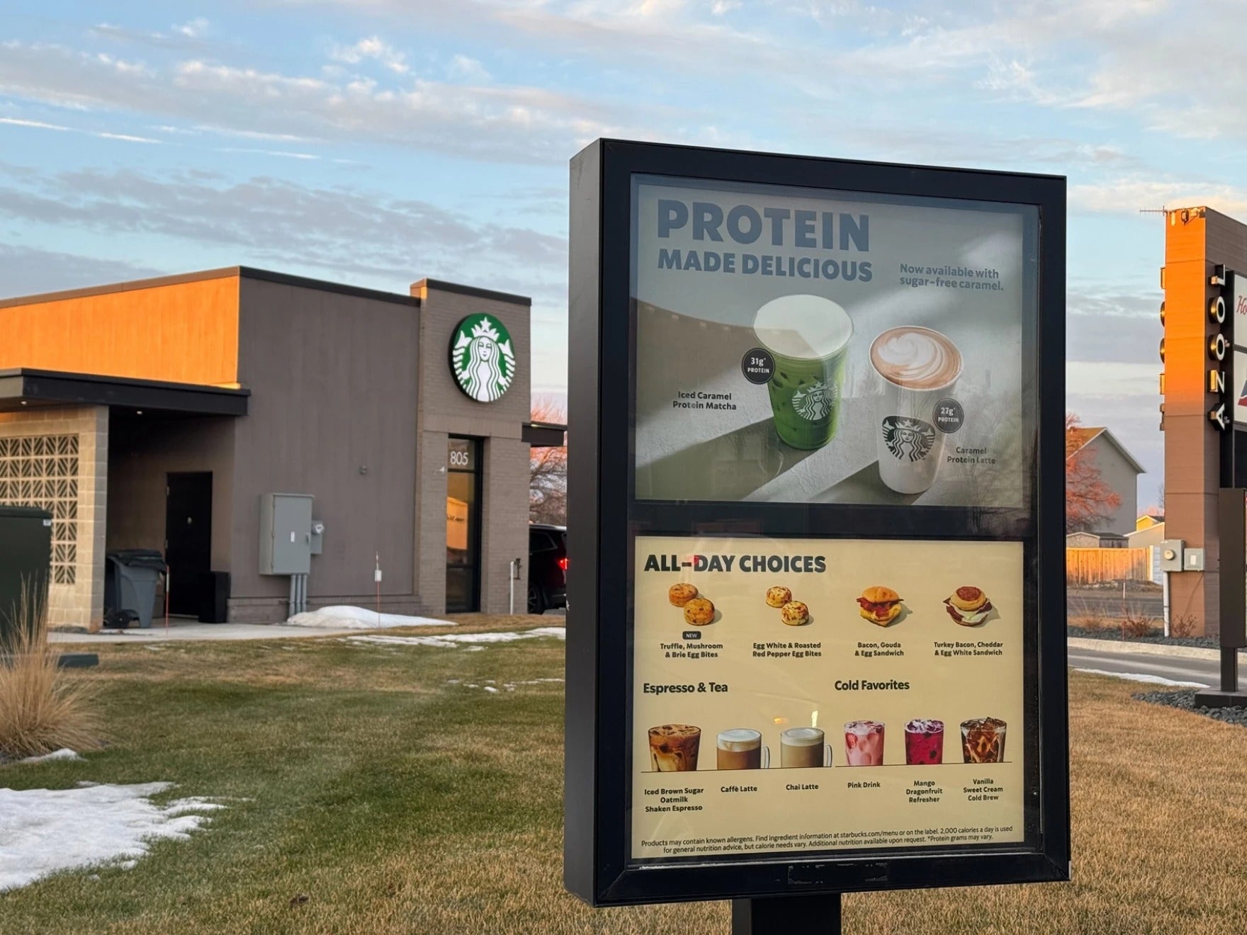 Starbucks drive-thru menu displays food and drink options, including espresso, tea, cold drinks, and protein beverages, with a Starbucks store in the background.