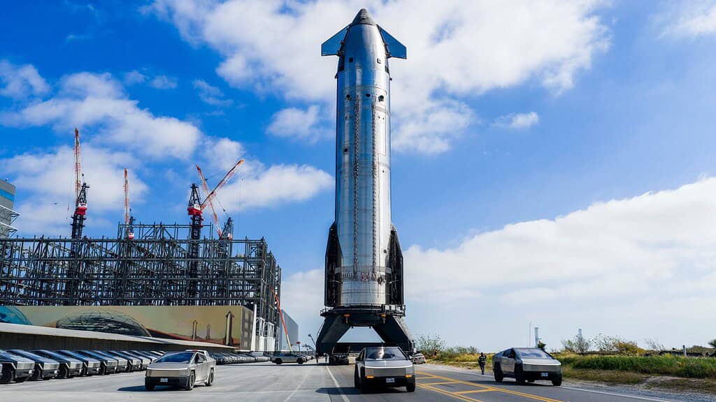 SpaceX's Starship rocket sitting on a launch pad in a lot behind a fleet of cybertrucks.