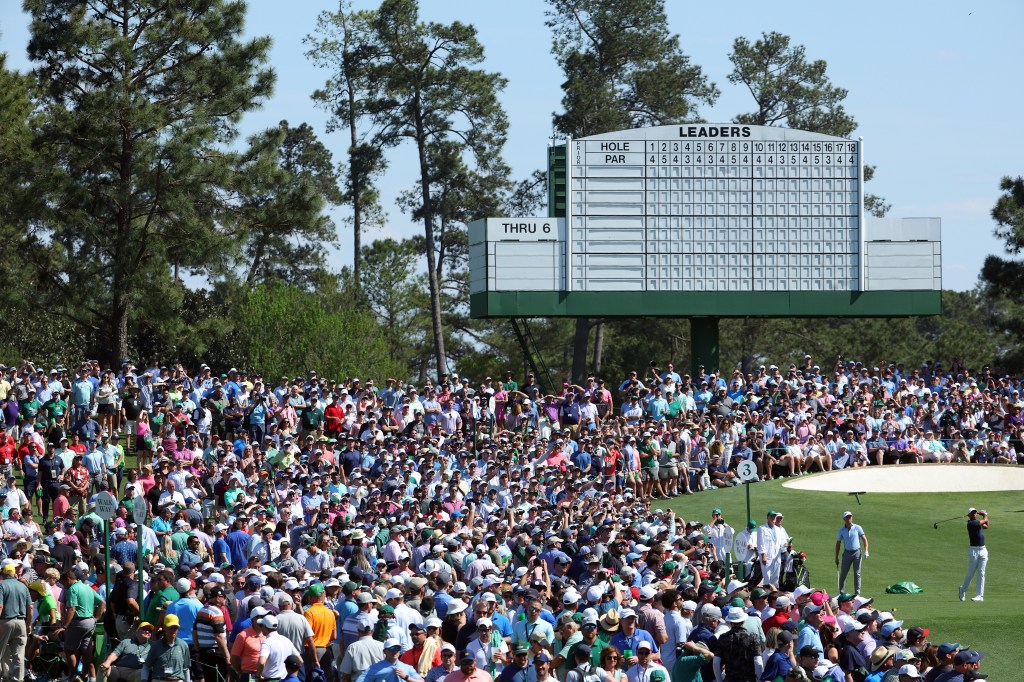 A golfer in a navy shirt and light-colored pants plays a shot at the Masters, watched by a large crowd and a leaderboard showing "LEADERS THRU 6."