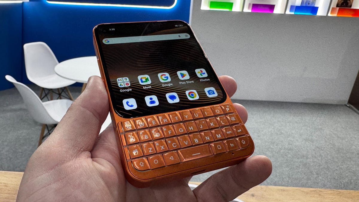 A hand holding a small-format orange phone with a full but compact QWERTY keyboard under a square phone screen with a modern Android interface.