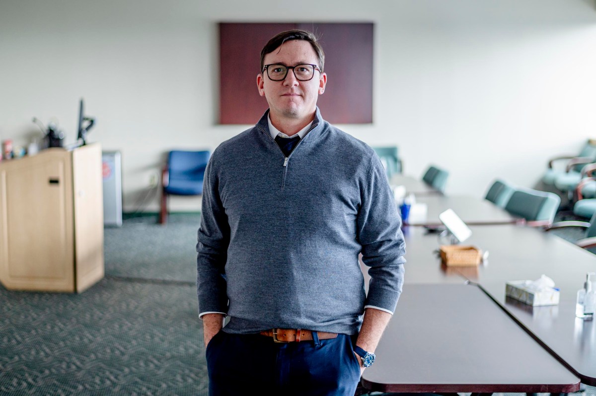 A man wearing glasses and a gray sweater stands with hands in pockets in a conference room with tables and chairs.