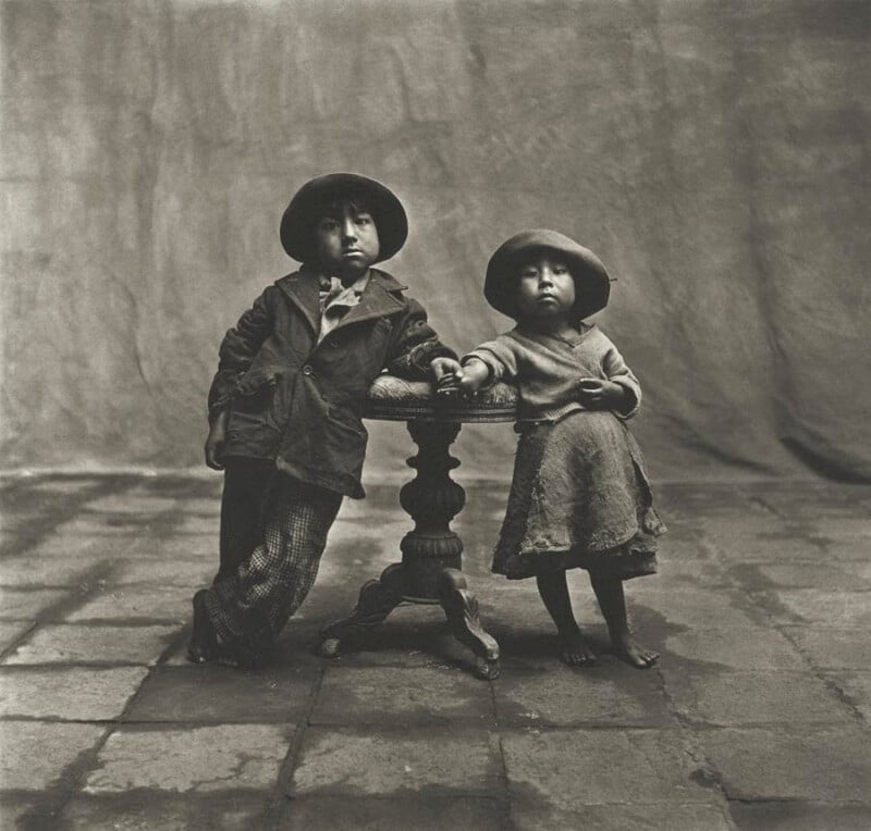Two young children in old-fashioned clothes and hats stand barefoot beside a small round table, looking at the camera. The background is draped fabric, and the floor is made of stone tiles. The photo is in black and white.