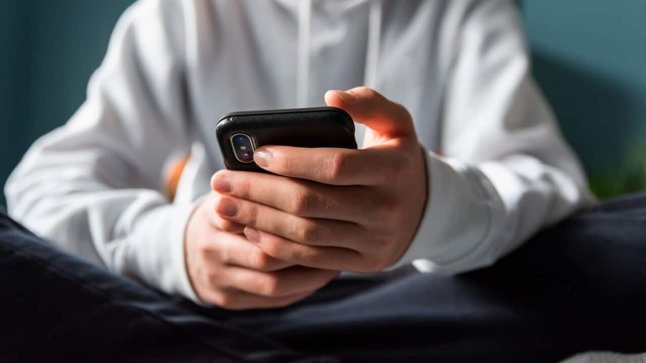 A boy wearing a white sweater, holding a black phone in his hands.