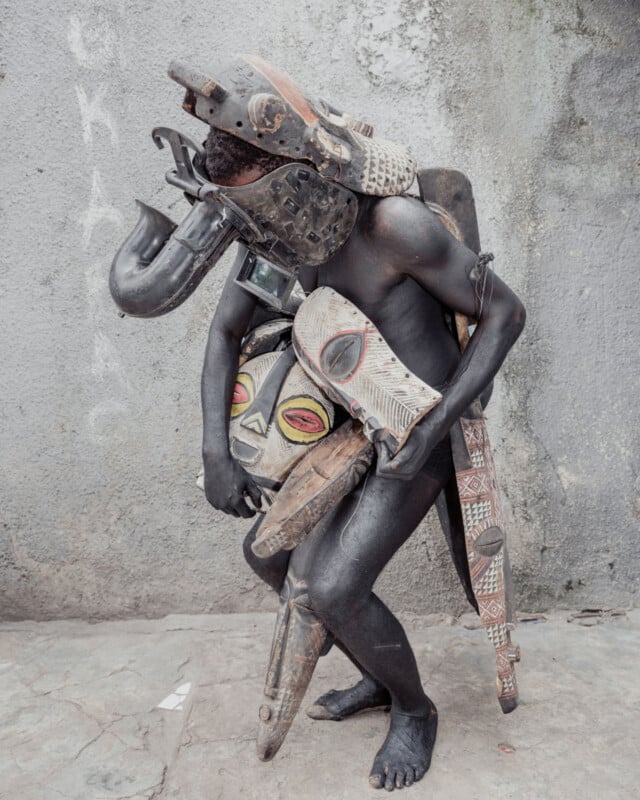 A person covered in black paint wears and carries multiple African tribal masks and artifacts, standing against a weathered gray wall on a concrete surface.