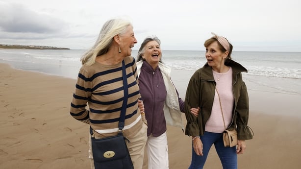 Three senior female friends walking along a beach enjoying time together, smiling and interacting in a relaxed, joyful moment. The image captures friendship, companionship and wellbeing, reflecting the pleasures of social connection and active, positive ageing in later life.