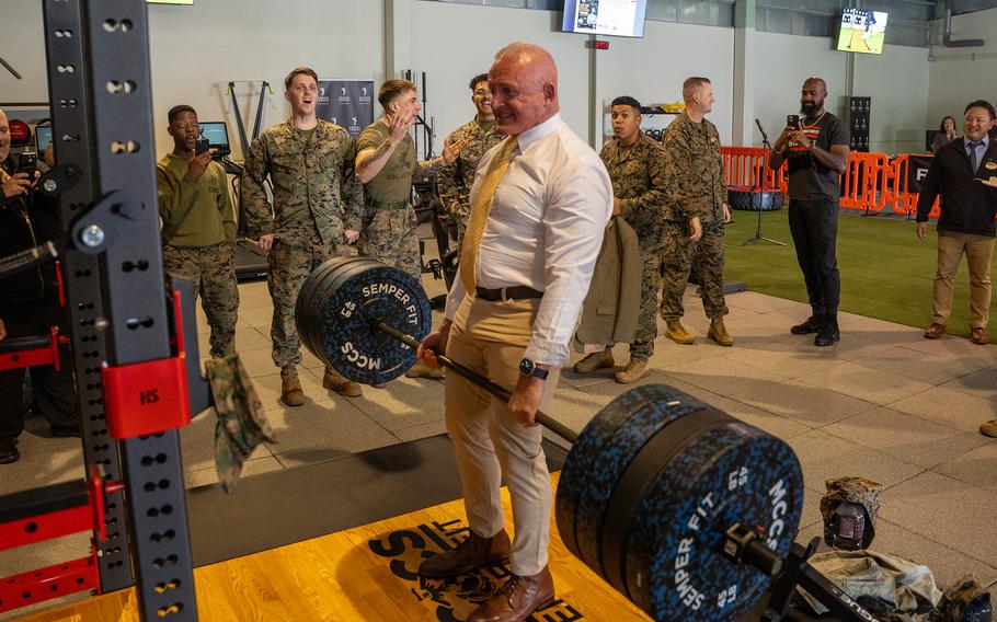 A man in a dress shirt, khakis and a tie performs a deadlift while Marines gather around and applaud during a strength training event at a base gym.